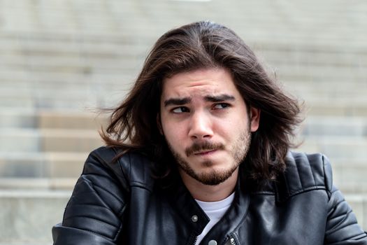 Portrait of an emotional bearded handsome man with long brown hair, dressed in trendy clothes, sitting on the steps and skeptical looking to the side, with a platform for advertising content. Handsome young posing on a copy space.