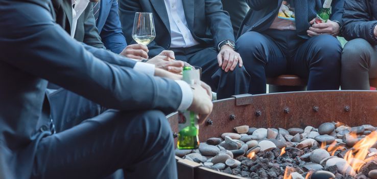 Group of Caucasian businessmen in formal dress gathering over the round fire pit after work in Chicago. Business people in formal dress with watches, leather shoes drinking beer bottle, wine glasses