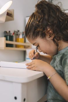 vertical photo of a little girl that writes concentrated at home with a color marker, she is drawing on the table in her room