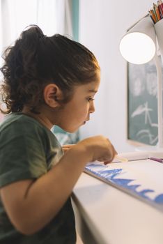 vertical photo of a little girl drawing concentrated at home with a color marker, she is drawing on the table in her room