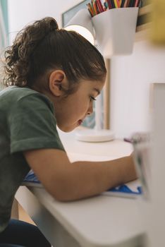 vertical photo of a little girl drawing concentrated at home with color markers, she is drawing on the table in her room