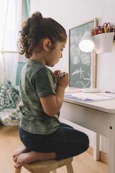 vertical photo of a little girl that draws concentrated at home with color markers, she is drawing on the table in her room