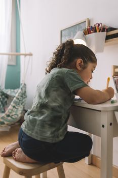 vertical photo of a little girl that draws concentrated at home with color markers, she is drawing on the desk in her room