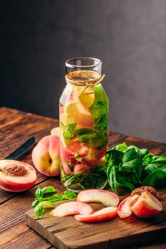 Bottle of Flavored Water with Sliced Peach and Basil Leaves. Knife and Ingredients on Cutting Board. Vertical Orientation.