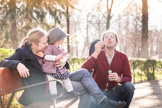 Young happy family with cheerful child having fun in park on sunny day. Father blowing soap bubbles.