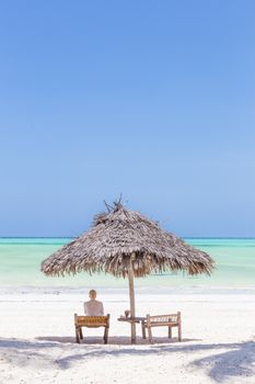 Woman reading on dack chair under wooden umbrella on white sandy tropical beach of Paje, Zanzibar, Tanzania. Copy space on blue sky.
