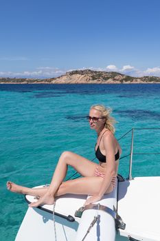 Woman in bikini tanning and relaxing on a summer sailin cruise, sitting on a luxury catamaran near picture perfect white sandy beach on Spargi island in Maddalena Archipelago, Sardinia, Italy.