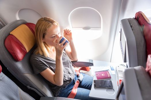 Woman on commercial passengers airplane during flight. Female traveler seated in passanger cabin drinking coffee. Sun shining trough airplane window.