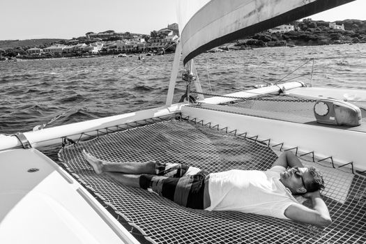 Sporty man relaxing, lying in hammock of a catamaran sailing boat on luxury nautic vacations near picture perfect Palau town, Sardinia, Italy. Black and white image.