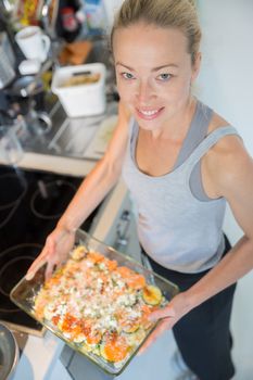 Smiling young healthy woman holding and proudly showing glass baking try with row vegetarian dish ingredients before placing it into oven. Healthy home-cooked everyday vegetarian food.