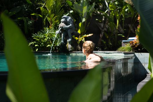 Sensual young woman relaxing in outdoor spa infinity swimming pool surrounded with lush tropical greenery of Ubud, Bali. Wellness, natural beauty and body care concept.