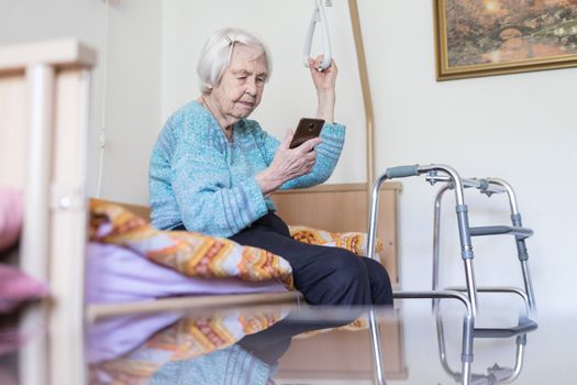 Elderly 96 years old woman reading phone message while sitting on medical bed supporting her by holder. Senior woman using wireless internet connection on smart phone.