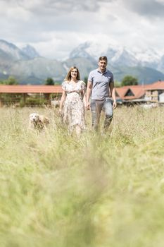 Young happy pregnant couple holding hands walking it's Golden retriever dog outdoors at countryside. Concept of pregnancy and family creation.