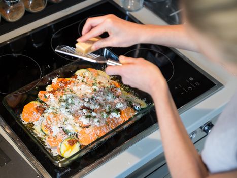 Female chef grinding parmesan cheese on row vegetarian dish ingredients in glass baking try before placing it into oven. Healthy home-cooked everyday vegetarian food.