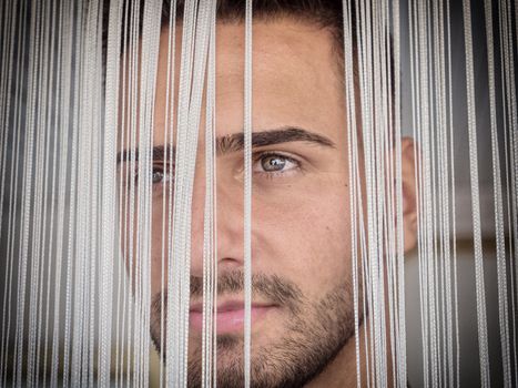 Headshot of handsome young man behind strings curtain, looking away to a side