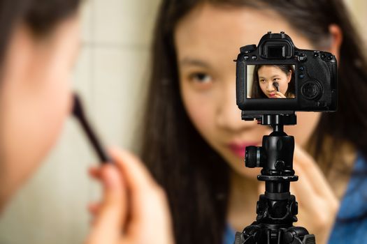 Focus on live view on camera on tripod, teenage girl  using cosmetics image on back screen with blurred scene in background. Teenage vlogger livestreaming show concept