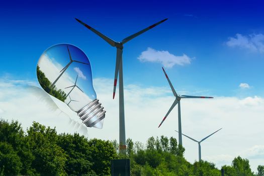 Light bulb with wind turbine inside. In the background blue sky and green trees with turbines. Conceptual image