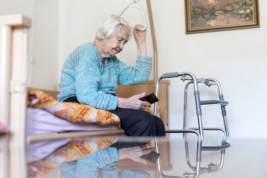 Elderly 96 years old woman reading phone message while sitting on medical bed supporting her by holder. Senior woman using wireless internet connection on smart phone.