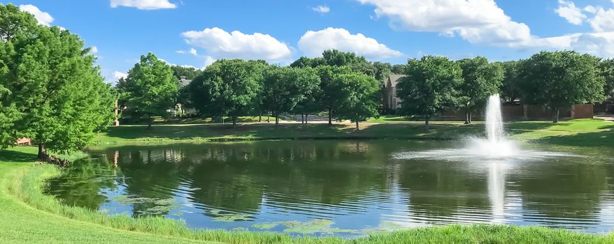 Panorama view beautiful pond with water fountain in small neighborhood North of Dallas, Texas, America. Lake house surrounding by matured trees, green grass lawn and cloud blue sky