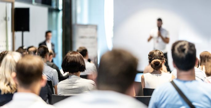 Business and entrepreneurship symposium. Speaker giving a talk at business meeting. Audience in conference hall. Rear view of unrecognized participant in audience.