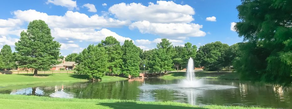 Panorama view beautiful pond with water fountain in small neighborhood North of Dallas, Texas, America. Lake house surrounding by matured trees, green grass lawn and cloud blue sky