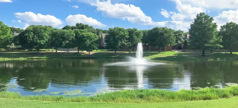 Beautiful pond with water fountain in small neighborhood North of Dallas, Texas, America. Lake house surrounding by matured trees, green grass lawn and cloud blue sky