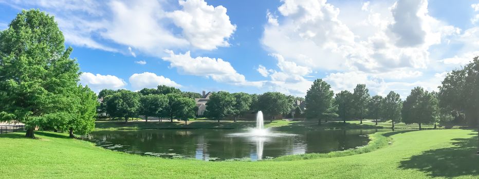 Panorama view beautiful pond with water fountain in small neighborhood North of Dallas, Texas, America. Lake house surrounding by matured trees, green grass lawn and cloud blue sky