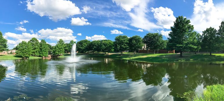 Panorama view beautiful pond with water fountain in small neighborhood North of Dallas, Texas, America. Lake house surrounding by matured trees, green grass lawn and cloud blue sky