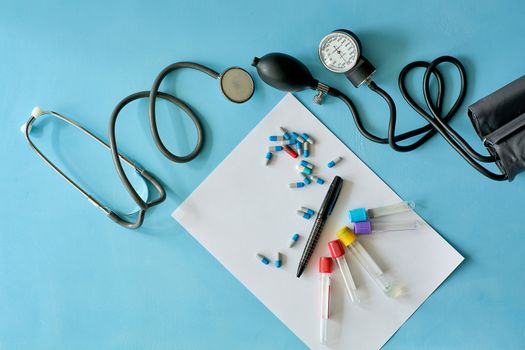 White sheet paper with black pen and colored different pills, capsules, stethoscope, sphygmomanometer, vacuum venipuncture test tubes on blue background with copy space for text. Medicine consept.