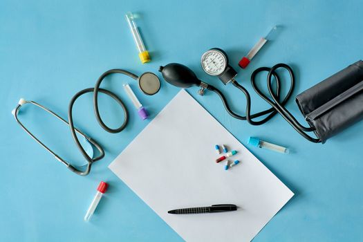 White sheet paper with black pen and colored different pills, capsules, stethoscope, sphygmomanometer, vacuum venipuncture test tubes on blue background with copy space for text. Medicine consept.