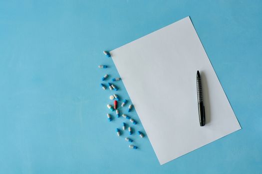 White sheet paper with black pen and colored different pills, capsules on blue background with copy space for text. Medicine consept. Print disign consept.