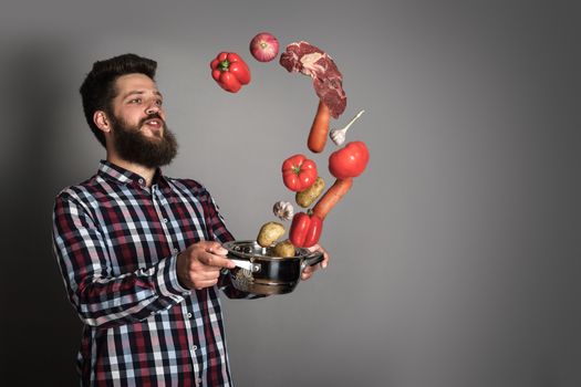 Cooking man concept, smiling bearded man in checked shirt, drop up meat and vegetables from a pan, studio shot on gray background