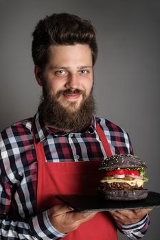 Male cook in red apron giving you fresh self made black burger and smiling