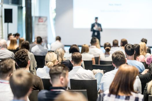 Business and entrepreneurship symposium. Speaker giving a talk at business meeting. Audience in conference hall. Rear view of unrecognized participant in audience.