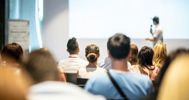 Business and entrepreneurship symposium. Speaker giving a talk at business meeting. Audience in conference hall. Rear view of unrecognized participant in audience.