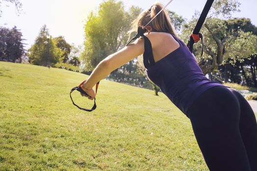 young beautiful blonde woman using suspension straps, exercise routine outdoors at the evening in a sunny park