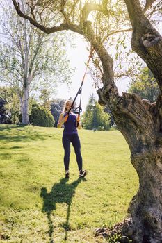 vertical photo of a beautiful young woman training outdoors under a tree, fitness girl workout with suspension equipment at the morning in a sunny park