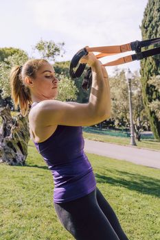 vertical photo of a strong young woman training biceps with suspension fitness straps, she is outdoors during functional workout
