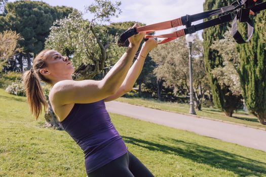 athletic young woman doing bicep curls with fitness suspension straps, during functional workout in a outdoor park