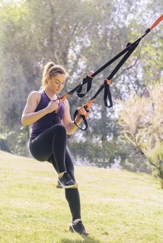 vertical photo of a strong young woman training with fitness suspension straps, during functional workout in a sunny outdoor park at the morning