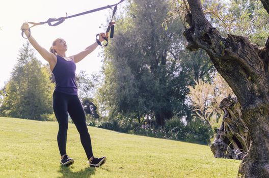 athletic young woman training with fitness suspension straps, during functional workout in a sunny outdoor park in the morning