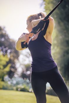 vertical photo of an athletic woman training with fitness straps, during functional workout in a sunny outdoor park in the morning
