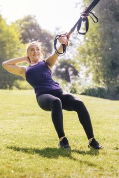 vertical photo of an athletic fitness woman that smiles training with suspension straps, during functional workout in a sunny outdoor park in the morning