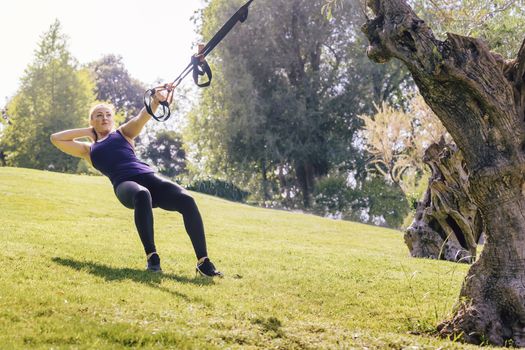 pretty fitness woman training with suspension straps, during functional workout in a sunny outdoor park in the morning