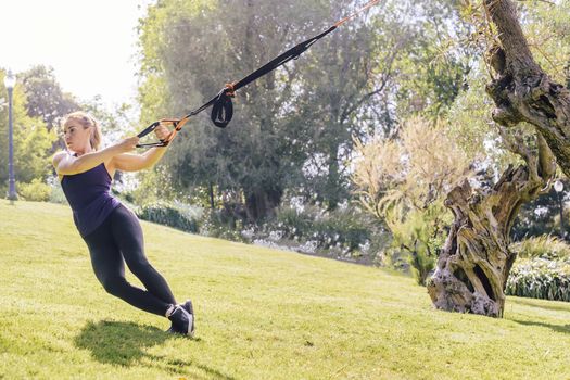 young beautiful blonde woman using suspension straps, functional workout in a sunny outdoor park in the morning