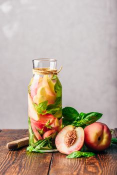 Bottle of Infused Water with Sliced Peach and Basil Leaves. Knife and Ingredients on Wooden Table. Vertical Orientation.