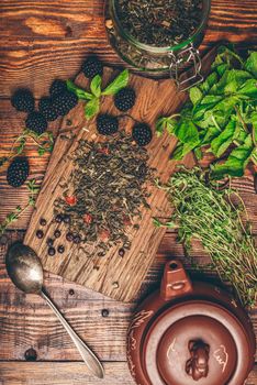 Heap of Dry Green Tea and Fresh Blackberries on Wooden Cutting Board. Bundles of Mint and Thyme Leaves. Clay Kettle. View from Above.