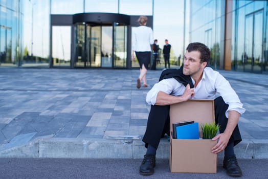 Fired business man sitting frustrated and upset on the street near office building with box of his belongings. He lost work