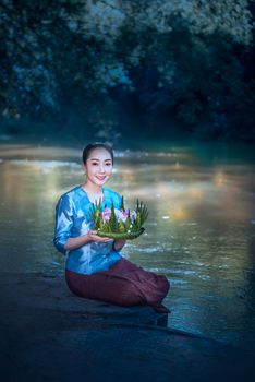 beautiful woman in traditional asian dresses holding vessel made of banana leaves and flowers sitting near river ( Loy Krathong Traditional Festival )