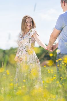 Young happy pregnant couple in love holding hands, relaxing in meadow. Concept of love, relationship, care, devotion, marriage, family creation, pregnancy, parenting
